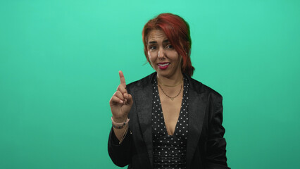 Obraz premium Woman pointing index finger upward in teal studio with solid green backdrop wearing polka dot dress and layered necklaces; firm caution.