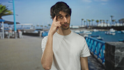 Fototapeta premium Young man in white tshirt raises finger to head and shows bare forearm on sunlit street overlooking boats; thoughtful insight.