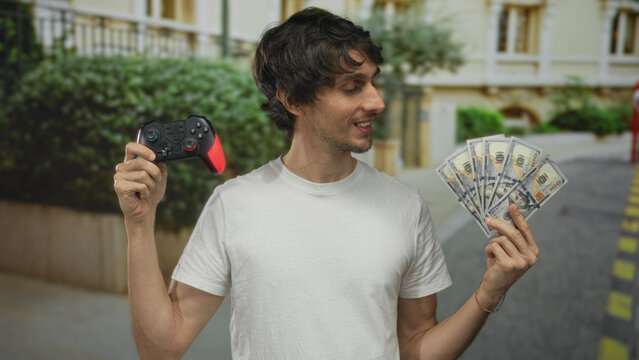 Man holding gaming controller in one hand and fanned cash with other hand on city street; winning thrill.