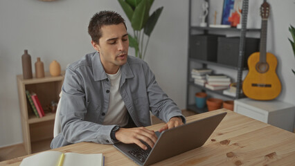 Young hispanic man working on laptop in cozy living room with guitar and bookshelf background showcasing a casual home environment with plants and decor elements.