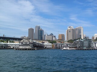 Fototapeta premium Sydney harbor cityscape with waterfront buildings and skyline under blue sky. Modern Australian urban scene.