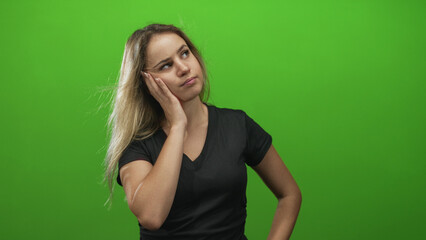 Young blonde woman touching cheek with hand and pointing finger upward in green studio; thoughtful curiosity.