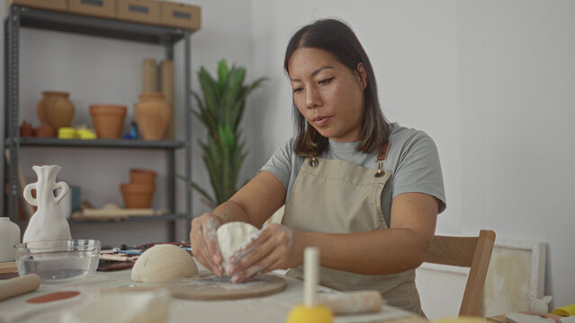 Woman shaping clay with hands in pottery studio workspace molding a bowl on table wearing apron with tools and ceramic pots on shelves; calm creativity.