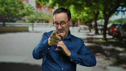 Man smiling in blue shirt gestures in sunny urban street with trees and blurred background suggesting cheerful outdoor setting with mature hispanic male in focus.
