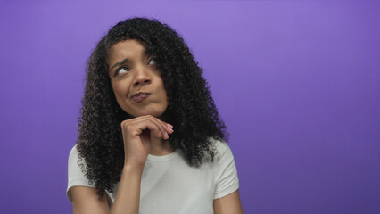 African american woman with curly hair looks upward with hand on chin in studio; thought doubt contemplation introspection.