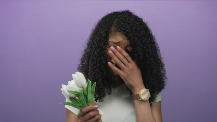 Woman with curly hair holding white tulip bouquet with hand pressed to face in purple studio; sadness.