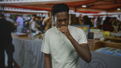 Obraz premium Man coughing into fist at a bustling street market stall surrounded by souvenir booths under canvas canopies; pain.