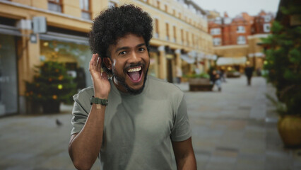 Man smiling on city street making gesture with thumb up and listening pose, surrounded by urban...