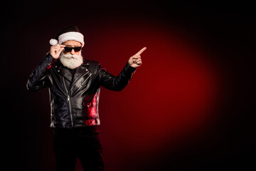 Cool Santa rocker in leather jacket with beard and sunglass points to right against red glow ready Christmas party and holiday celebration