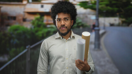 African american man holding blueprints on a city street, captured outdoors with buildings and...