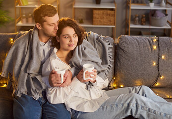 Loving young family at home feel comfortable under warm blankets on sofa, close happy Valentine day couple date, affectionate people in love, hug, romantic holiday room garland lights decoration