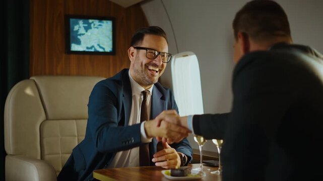 Two smiling businessmen in formal suits finalizing a deal and shaking hands while flying first class in a private jet, celebrating their successful partnership with champagne and a meal
