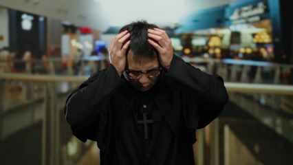 Obraz premium Priest wearing black robe and glasses in a mall expresses stress while holding head with both hands in front of blurred indoor shopping center background