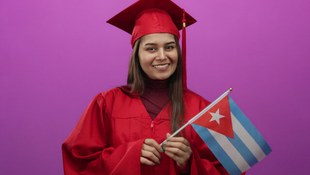 Woman in red graduation gown holding cuban flag on pink background, symbolizing academic achievement and cultural pride.