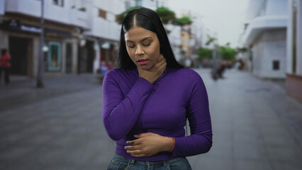 Fototapeta premium Young latin woman with long black hair clutching neck on city street wearing purple top and blue jeans; discomfort.