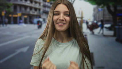 Fototapeta premium Teenager girl in green shirt clenches fists and smiles on urban street with blurred buildings; determination and energy.