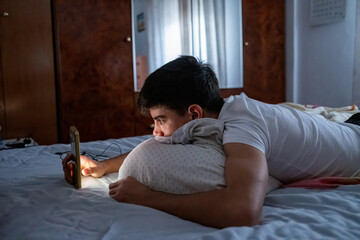 Teenage boy lies on bed in a dark room illuminated by smartphone light, browsing or texting before sleep. Concept of digital night habits. © Manuel