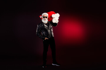 Cool Santa rocker in leather jacket holds a red gift bag wearing Santa hat and sunglass for Christmas fashion shoot