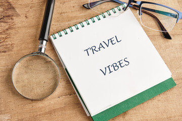 A notebook that says Travel Vibes sits on a wooden table, along with a magnifying glass and blue framed glasses, suggesting travel planning