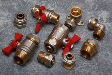 Brass and metal plumbing fittings and valves with red handles arranged on a gray background, showcasing various types of pipe connectors and adapters for water and gas systems.
