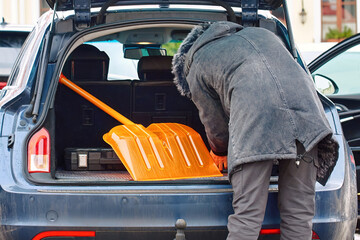 Man organizing snow removal tools in car trunk during cold season, safety preparation for winter driving and roadside emergencies. Driver retrieving snow shovel from vehicle boot in snowy city area