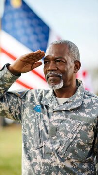 Mature man in camouflage military uniform saluting, standing in front of American flag outdoors, wearing service patch and gray hair