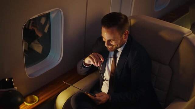 Confident middle-aged businessman working on a tablet computer and smiling during a comfortable flight in the first class cabin of a private jet, enjoying a luxurious business trip