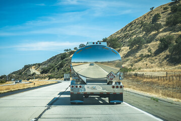 Fuel tanker truck driving on California highway through dry hills, bright sunny day, mirror reflection of the road in the shiny metal tank, travel and transportation scene, perspective view, minimalis