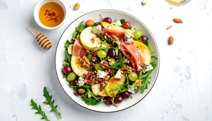 Overhead shot of a fresh salad in a white bowl, showcasing pear slices, grapes, prosciutto, nuts, and blue cheese. Honey and a honey dipper are nearby