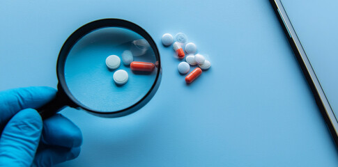 Person in blue gloves holding a magnifying glass over pills and capsules on a blue background