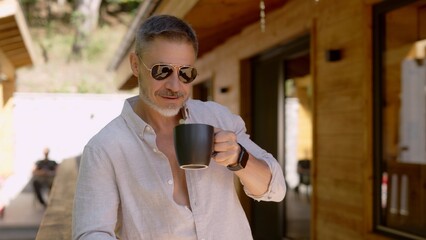 Mid adult man drinking morning coffee on terrace at home wearing sunglasses. Vacation in the mountain cabin house. Freedom, lifestyle and getaway.