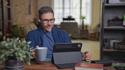 Middle aged man in glasses sitting at desk in home office remote working on digital tablet,...