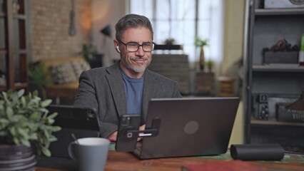 Happy confident businessman working with laptop and digital technology at home office desk, smiling. Middle aged man, male entrepreneur remote managing business. Telecommunication.