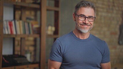 Portrait of happy casual middle aged man at home in loft room. Senior male with grey hair looking at camera in eyeglasses, smiling. Books and typewriter in background, smart older guy.