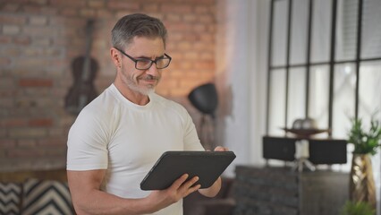 Middle aged man using digital table at home in loft living room. Happy, sporty, active senior male with gray hair and glasses reading social media, working and smiling.