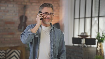 Happy middle aged man talking on phone call at home. Mid adult older male in glasses smiling, communicating using mobile. Telecommunication, entrepreneurship.