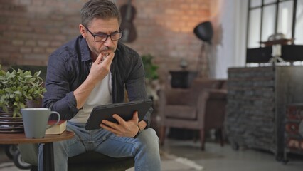 Senior businessman sitting in armchair in living room remote working in home office with digital tablet. Middle aged man, smiling reading. Entrepreneur managing business, thinking.