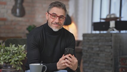 Senior businessman sitting in armchair in living room remote working in home office with phone. Portrait of happy, confident middle aged man, smiling. Entrepreneur managing business.