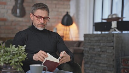 Middle aged man sitting in living room reading book wearing turtleneck pullover and eyeglasses. Smart looking mid adult male enjoying leisure and free time at home.