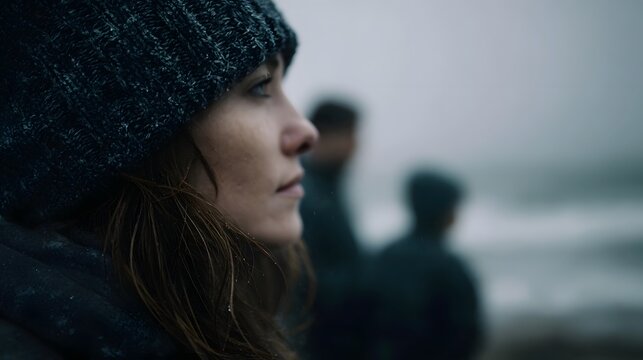 A woman in a beanie looks out at a misty sea on a cloudy day with blurred figures behind her - Powered by Adobe