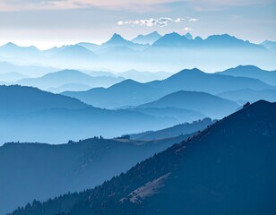 Majestic mountain range bathed in layers of atmospheric mist, creating a gradient of blue hues. Sky hints at a sunrise or sunset