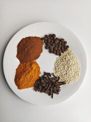Top view of assorted spices on white plate: ground nutmeg, turmeric, sesame seeds, whole cloves and peppercorns, minimal kitchen style and warm earthy tones