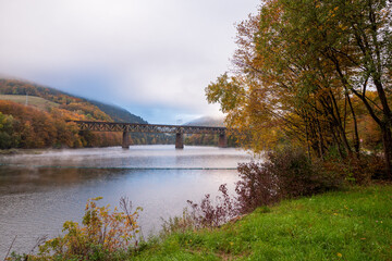 Fototapeta premium Railway bridge over Ružín reservoir in Slovakia during autumn. Calm lake, colorful forest, morning fog and peaceful nature landscape in mountain valley.