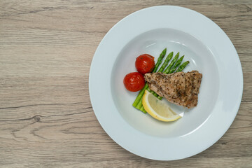 Red snapper steak, asparagus, tomatoes, and lemon on a white  plate on wooden background. Table top view.