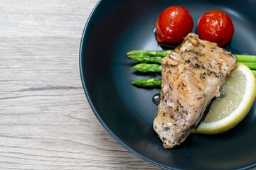 Red snapper steak, asparagus, tomatoes, and lemon on a black plate on wooden background. Table top view.