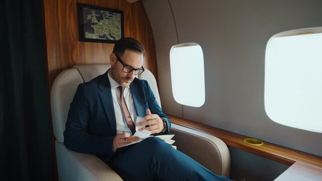 Focused businessman wearing a suit and glasses writing down notes in a personal notebook while sitting comfortably in a luxury private jet on a business trip to another country