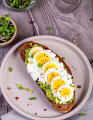 A slice of bread topped with mashed avocado, a creamy spread, sliced hard-boiled eggs, and microgreens. The dish is presented on a plate, with a bowl of additional microgreens nearby.