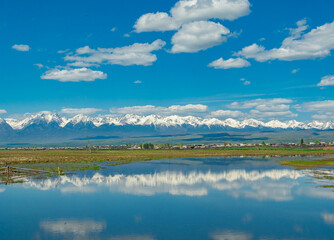 Majestic snow-capped mountains reflect in serene lake under bright blue sky with fluffy clouds in summer
