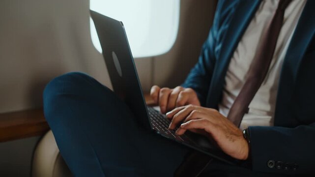 Successful entrepreneur wearing a suit works on his laptop while traveling in first class on a private jet, managing his business remotely during an important corporate trip for a meeting