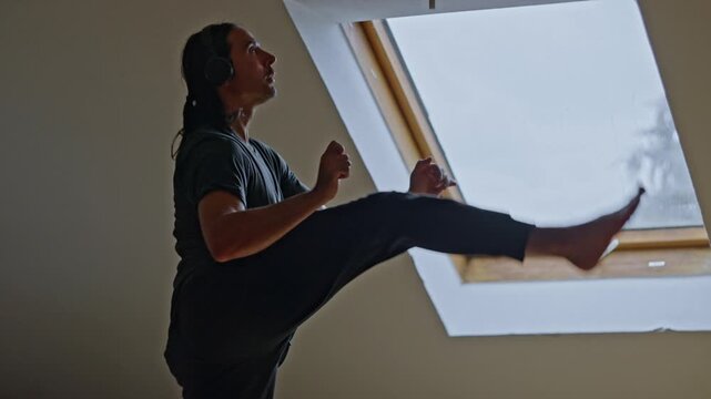 energetic martial arts workout near skylight window, high kick exercise session with music in spacious loft, dynamic cardio and martial rhythm training performed by man in airy loft